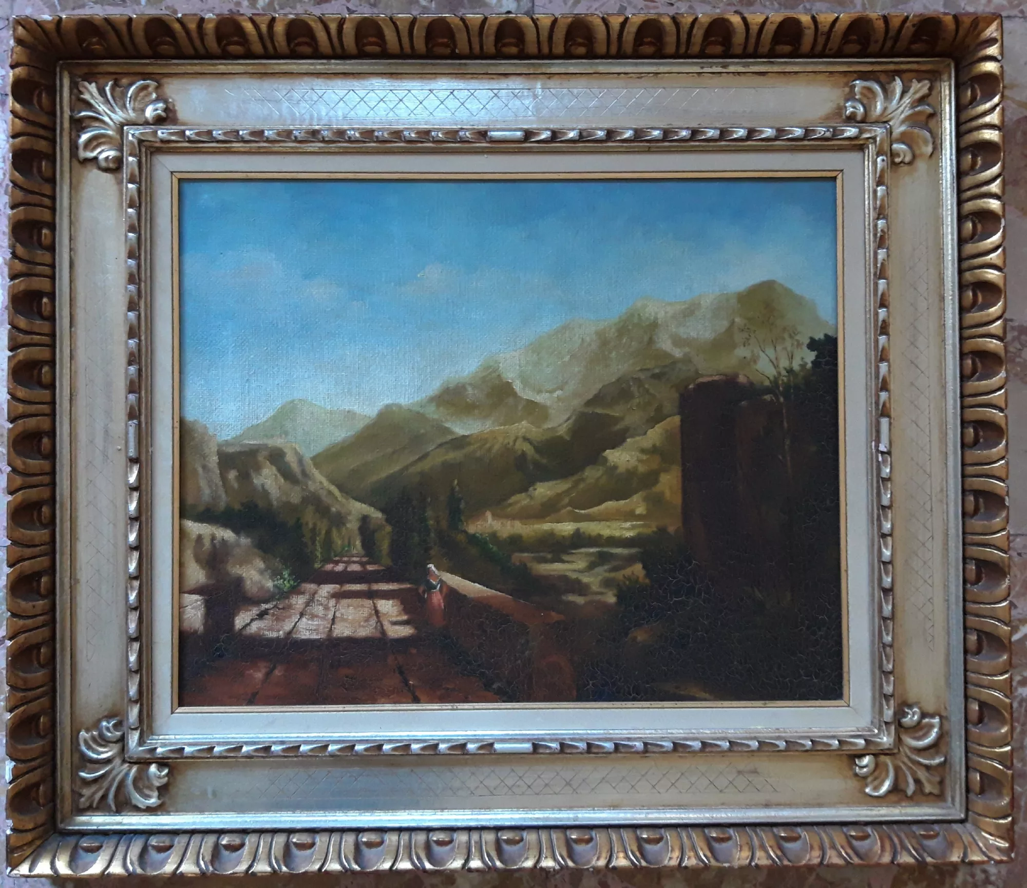 Paesaggio-montano-con-craquelere-Aly-Mohsen "Quadro di Ay Mohsen realizzato ad Olio con pennello. Il titolo è Paesaggio montano con craquelure e rientra nella categoria Paesaggi. In Paesaggio montano con craquelure, Aly Mohsen propone una visione ampia e silenziosa del paesaggio montano, in cui una strada lastricata accompagna lo sguardo verso un orizzonte di colline e montagne immerse in una luce morbida e diffusa. La presenza umana, piccola e discreta, introduce una misura di scala e rafforza il dialogo tra uomo e natura, suggerendo un’idea di attraversamento e contemplazione. La composizione è costruita su una solida profondità prospettica, articolata in piani successivi che si susseguono con naturalezza fino alle cime lontane. I toni caldi della terra e i verdi smorzati delle pendici montane si fondono gradualmente con l’azzurro del cielo, creando un’atmosfera di quiete sospesa e di equilibrio naturale. La pennellata è misurata e narrativa, capace di restituire la solidità del terreno, la morbidezza delle colline e l’ampiezza dello spazio. In modo consapevole, Aly Mohsen ha ricreato una craquelure pittorica controllata, conferendo alla superficie del dipinto l’aspetto di un’opera antica. Questa scelta non è puramente estetica, ma testimonia la padronanza tecnica dell’artista e la sua capacità di dialogare con i procedimenti storici della pittura, in particolare con la tradizione del paesaggio classico europeo. La luce, diffusa e priva di drammatizzazioni, accompagna lo sguardo lungo il percorso visivo, favorendo una lettura lenta e contemplativa dell’opera. L’insieme restituisce una sensazione di tempo rallentato e di armonia tra natura e presenza umana. La cornice dorata, ricca e decorativa, valorizza ulteriormente il dipinto, rafforzandone il carattere classico e accompagnando la composizione con equilibrio ed eleganza. Paesaggio montano con craquelure si inserisce nella produzione paesaggistica di Aly Mohsen come un’opera di grande equilibrio formale e profondità visiva, capace di coniugare memoria storica, competenza tecnica e sensibilità poetica."