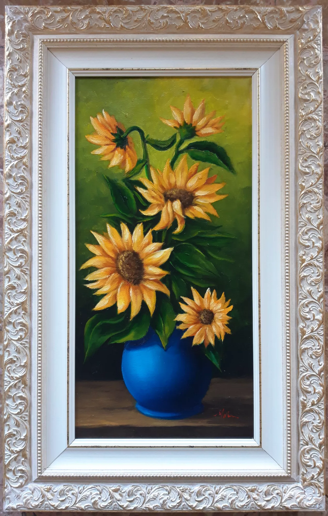 Vaso-con-girasoli-Aly-Mohsen "Quadro di Ay Mohsen realizzato ad Olio con pennello. Il titolo è Vaso con girasoli e rientra nella categoria Natura morta. Vaso con girasoli di Aly Mohsen è una natura morta floreale che si distingue per la chiarezza compositiva e per l’intensa relazione cromatica tra soggetto e sfondo. L’opera si inserisce nella tradizione della pittura figurativa, affrontando il tema floreale con un linguaggio sobrio e luminoso, attento alla resa della luce e alla vitalità delle forme naturali. La composizione raffigura un bouquet di girasoli disposto con naturalezza all’interno di un vaso blu intenso, collocato su un piano orizzontale. Il vaso costituisce il fulcro visivo dell’opera e crea un deciso contrasto cromatico con i gialli caldi dei petali e con i verdi profondi delle foglie. I fiori, rappresentati in diverse inclinazioni e stadi di apertura, introducono un ritmo dinamico che conferisce equilibrio e movimento all’insieme. Lo sfondo verde, uniforme e leggermente sfumato, avvolge la composizione senza interferenze narrative, mettendo in risalto la luminosità dei girasoli e rafforzando la percezione spaziale. La luce, diffusa e controllata, modella i petali e le foglie con gradualità, evidenziandone la struttura materica e la volumetria. La pittura, realizzata a pennello, mostra una tecnica sicura e una particolare attenzione alla resa delle superfici e dei passaggi tonali. L’opera testimonia l’interesse di Aly Mohsen per il soggetto naturale come occasione di studio formale e cromatico, nel solco della tradizione della natura morta, reinterpretata con sensibilità contemporanea. Il dipinto è completato da una cornice di gusto classico, caratterizzata da un’ampia modanatura decorata in tonalità chiare con motivi ornamentali in rilievo, che incornicia l’opera con equilibrio ed eleganza, valorizzandone la verticalità e rafforzandone la presentazione di carattere museale."