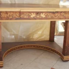 "Opera di Aly Mohsen realizzata con la tecnica dell'intarsio. Il titolo è Scrivania delle chimere e rientra nella categoria Ebanisteria Monumentale. La Scrivania delle Chimere è un’opera di ebanisteria artistica monumentale di Aly Mohsen, concepita come pezzo unico e realizzata secondo i principi della grande tradizione europea dell’alta falegnameria d’arte. L’opera si colloca al confine tra scultura funzionale e manufatto simbolico, integrando architettura, decorazione e iconografia in una struttura coerente e unitaria. Il piano è realizzato in radica di noce selezionata, caratterizzata da una venatura complessa e simmetrica, valorizzata da una finitura accurata che ne esalta la profondità cromatica e la qualità materica. La superficie è incorniciata da un intarsio policromo composto da acero bianco, ebano e padouk, disposto secondo un disegno continuo di gusto classico, eseguito con elevata precisione tecnica. La struttura portante è in mogano massello, utilizzato per le gambe, le fasce e gli elementi architettonici principali. Le superfici verticali sono arricchite da intarsi in acero bianco, interamente realizzati a mano, con motivi ornamentali e figurativi che dialogano con la struttura senza sovraccaricarla. La base inferiore curva e il collegamento tra le gambe contribuiscono alla stabilità visiva e strutturale dell’insieme. Elemento distintivo dell’opera è la presenza delle chimere alate intarsiate lungo le fasce laterali. Queste figure assumono un valore iconografico e simbolico, richiamando temi di protezione, forza e trasformazione, ricorrenti nella ricerca artistica di Mohsen. L’inserimento delle chimere non è puramente decorativo, ma parte integrante della concezione narrativa e formale dell’opera. La scrivania è concepita come opera d’arte funzionale, in cui l’uso pratico è subordinato a una visione artistica complessiva. La qualità dei materiali, la lavorazione manuale e l’unità tra struttura e decorazione ne fanno un manufatto destinato a contesti rappresentativi, in cui possa essere letto e osservato come oggetto artistico autonomo."
