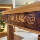 "Opera di Aly Mohsen realizzata con la tecnica dell'intarsio. Il titolo è Scrivania delle chimere e rientra nella categoria Ebanisteria Monumentale. La Scrivania delle Chimere è un’opera di ebanisteria artistica monumentale di Aly Mohsen, concepita come pezzo unico e realizzata secondo i principi della grande tradizione europea dell’alta falegnameria d’arte. L’opera si colloca al confine tra scultura funzionale e manufatto simbolico, integrando architettura, decorazione e iconografia in una struttura coerente e unitaria. Il piano è realizzato in radica di noce selezionata, caratterizzata da una venatura complessa e simmetrica, valorizzata da una finitura accurata che ne esalta la profondità cromatica e la qualità materica. La superficie è incorniciata da un intarsio policromo composto da acero bianco, ebano e padouk, disposto secondo un disegno continuo di gusto classico, eseguito con elevata precisione tecnica. La struttura portante è in mogano massello, utilizzato per le gambe, le fasce e gli elementi architettonici principali. Le superfici verticali sono arricchite da intarsi in acero bianco, interamente realizzati a mano, con motivi ornamentali e figurativi che dialogano con la struttura senza sovraccaricarla. La base inferiore curva e il collegamento tra le gambe contribuiscono alla stabilità visiva e strutturale dell’insieme. Elemento distintivo dell’opera è la presenza delle chimere alate intarsiate lungo le fasce laterali. Queste figure assumono un valore iconografico e simbolico, richiamando temi di protezione, forza e trasformazione, ricorrenti nella ricerca artistica di Mohsen. L’inserimento delle chimere non è puramente decorativo, ma parte integrante della concezione narrativa e formale dell’opera. La scrivania è concepita come opera d’arte funzionale, in cui l’uso pratico è subordinato a una visione artistica complessiva. La qualità dei materiali, la lavorazione manuale e l’unità tra struttura e decorazione ne fanno un manufatto destinato a contesti rappresentativi, in cui possa essere letto e osservato come oggetto artistico autonomo."