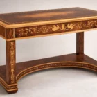 "Artwork by Aly Mohsen executed in the marquetry technique. The title is Scrivania delle chimere (Desk of the Chimeras – original Italian title) and it belongs to the category Monumental Cabinetmaking. The Scrivania delle Chimere is a monumental work of artistic cabinetmaking by Aly Mohsen, conceived as a unique, one-of-a-kind piece and executed in accordance with the traditions of high European fine woodworking. The work occupies a space between functional sculpture and symbolic artifact, integrating structure, decoration, and iconography into a unified artistic whole. The desktop is crafted from carefully selected walnut burl, distinguished by its complex, symmetrical grain pattern, enhanced through a refined finish that emphasizes both chromatic depth and material presence. The surface is framed by a polychrome marquetry border composed of white maple, ebony, and padauk, arranged in a continuous classical motif and executed with a high degree of technical precision. The supporting structure is made of solid mahogany, used for the legs, aprons, and primary architectural elements. Vertical surfaces are enriched with hand-cut white maple inlays featuring ornamental and figurative motifs that engage in a balanced dialogue with the overall structure. The curved lower base and the connecting elements between the legs contribute to both the visual stability and the structural coherence of the piece. A defining feature of the work is the presence of winged chimeras inlaid along the side panels. These figures carry an iconographic and symbolic significance, evoking themes of protection, strength, and transformation—recurring elements within Mohsen’s artistic research. Their inclusion is not merely decorative, but integral to the conceptual and narrative framework of the desk. The desk is conceived as a functional artwork, in which practical use is subordinate to an overarching artistic vision. The quality of the materials, the entirely hand-executed craftsmanship, and the unity between structure and decoration establish the piece as an autonomous artistic object, intended for representational settings where it can be experienced and interpreted as a work of art in its own right."