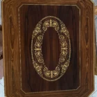 "Opera di Ay Mohsen realizzata con la tecnica dell'intarsio. Il titolo è Scrivania degli angioletti e rientra nella categoria Ebanisteria Monumentale. Opera unica di ebanisteria artistica contemporanea, la scrivania si colloca nell’ambito delle arti applicate come esempio di sintesi tra funzione e ricerca formale nella produzione di Aly Mohsen. Il manufatto è concepito come oggetto autonomo, realizzato secondo tecniche tradizionali di lavorazione del legno e intarsio manuale, con un’impostazione monumentale e misurata. La struttura è eseguita in noce nazionale, selezionato per la stabilità strutturale e la profondità cromatica, accostato a palissandro dalle tonalità più calde e brunite, impiegato per definire superfici e volumi. Il piano ottagonale presenta un intarsio centrale in acero bianco, disposto secondo un motivo ornamentale di matrice classica, eseguito interamente a mano e inserito entro una cornice perimetrale che ordina visivamente la superficie. Le fasce laterali e le gambe sagomate sono arricchite da ulteriori intarsi in acero, con figure allegoriche e motivi floreali sviluppati in sequenza ritmica. La decorazione si integra alla struttura senza sovrapporsi ad essa, contribuendo alla leggibilità dell’insieme e alla continuità tra elemento architettonico e apparato ornamentale. L’uso combinato delle essenze lignee genera un equilibrio cromatico controllato, mentre la finitura, sobria e uniforme, è finalizzata a valorizzare la venatura naturale dei materiali e la precisione dell’intarsio, senza effetti di enfatizzazione superficiale. La scrivania è concepita come opera d’arte funzionale, destinata a contesti di rappresentanza, spazi espositivi o ambienti dedicati alle arti applicate, come testimonianza di un sapere ebanistico fondato sulla manualità, sul progetto unitario e sulla continuità con la tradizione storica europea."