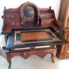 "Opera di Ay Mohsen realizzato con la tecnica dell'intarsio. Il titolo è Scrittoio della musica e rientra nella categoria Ebanisteria Monumentale. Questo straordinario Scrittoio della Musica, firmato Aly Mohsen, rappresenta uno dei vertici assoluti della sua produzione di ebanisteria artistica, in cui pittura, scultura e intarsio ligneo convivono in un unico oggetto funzionale e fortemente simbolico. L’opera si configura come un mobile scultoreo d’arte, concepito non come semplice arredo ma come opera autonoma, capace di dialogare con la tradizione delle arti applicate europee di alto livello. La struttura architettonica richiama con coerenza il linguaggio neoclassico e Impero, attraverso volumi pieni, una raffinata alzata centrale ad arco e colonne tornite coronate da capitelli in bronzo brunito, elementi che conferiscono solennità, ritmo e autorevolezza all’insieme. Il dialogo tra rigore formale e ricchezza decorativa costituisce uno dei tratti distintivi dell’opera. I pannelli frontali, laterali e posteriori sono impreziositi da intarsi figurativi a tema musicale e ornamentale: strumenti, spartiti, ghirlande e una raffinata composizione floreale in vaso, simboli universali di armonia, creatività e bellezza senza tempo. Particolarmente significativa è la decorazione del retro, concepita come un’opera completa e autonoma, scelta rara e consapevole che ribadisce la natura artistica totale dello scrittoio. Dal punto di vista materico, l’opera è realizzata attraverso una sapiente combinazione di radica di noce e radica di thuya, selezionate per la profondità cromatica e la vivacità delle venature. Acero, ebano e padouk sono impiegati per cornici, filettature e contrasti cromatici, mentre il pero definisce con eleganza la cornice dell’intarsio posteriore, introducendo una tonalità calda e luminosa che armonizza l’intera composizione. Il piano di scrittura estraibile, scandito da campiture scure e filetti chiari, incornicia un riquadro centrale intarsiato che funge da fulcro visivo e funzionale. I numerosi cassetti, perfettamente integrati nella struttura, testimoniano l’elevatissimo livello tecnico dell’artista e la sua capacità di coniugare complessità costruttiva ed equilibrio formale. Interamente realizzato a mano, senza alcun ricorso a processi industriali, lo Scrittoio della Musica si impone come testimonianza dell’alta ebanisteria artistica europea contemporanea, in cui si rinnova consapevolmente la grande tradizione delle arti applicate storiche. L’opera si colloca idealmente tra arte applicata, scultura funzionale e patrimonio culturale, risultando idonea a contesti museali, collezioni storiche e ambienti di rappresentanza di alto profilo culturale."