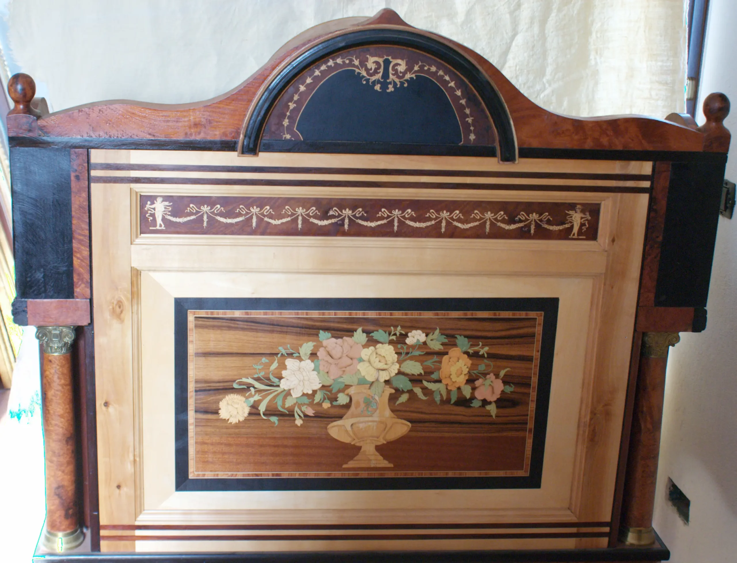 "Artwork by Aly Mohsen executed in the marquetry technique. The title is Scrittoio della musica (Music Writing Desk – original Italian title) and it belongs to the category Monumental Cabinetmaking. Scrittoio della Musica is an extraordinary artistic writing desk by Aly Mohsen and stands as one of the highest achievements of his artistic cabinetmaking. In this work, painting, sculpture, and marquetry converge into a single functional yet profoundly symbolic object. Conceived not as mere furniture, the desk presents itself as an autonomous sculptural artwork, firmly rooted in the tradition of European high decorative arts. The architectural structure clearly references Neoclassical and Empire styles, articulated through solid volumes, a refined central arched superstructure, and turned columns crowned with burnished bronze capitals. These elements lend the piece rhythm, solemnity, and a strong sense of formal authority. The balance between architectural rigor and decorative richness is a defining characteristic of the work. The front, side, and rear panels are enriched with figurative marquetry inspired by music and ornamentation: musical instruments, scores, garlands, and an elegant floral composition in a vase, alluding to harmony, creativity, and the timeless nature of art. Of particular significance is the fully decorated back panel, conceived as an independent artwork—an intentional and rare choice that reinforces the sculptural and artistic identity of the desk. From a material perspective, the piece is crafted using a refined combination of walnut burl and thuya burl, selected for their chromatic depth and vibrant grain patterns. Maple, ebony, and padauk are employed for frames, stringing, and contrasting elements, while pearwood defines the rear marquetry frame, introducing a warm and luminous tonal balance. The pull-out writing surface, structured with dark fields and light stringing, frames a central inlaid panel that serves as both a visual and functional focal point. The numerous drawers, seamlessly integrated into the structure, demonstrate Mohsen’s exceptional technical mastery and his ability to reconcile structural complexity with formal clarity. Entirely handcrafted, without the use of industrial processes, Scrittoio della Musica stands as a significant example of contemporary European artistic cabinetmaking, consciously renewing the great tradition of historic decorative and applied arts. The work occupies a position at the intersection of applied art, functional sculpture, and cultural heritage, making it particularly suited to museum contexts, historical collections, and high-profile cultural environments."