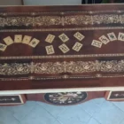 "Opera di Ay Mohsen realizzata con la tecnica dell'intarsio. Il titolo è Scrivania del poker e le tre carte e rientra nella categoria Ebanisteria Monumentale. Questa imponente scrivania monumentale rappresenta una delle opere più concettualmente audaci, raffinate e politicamente consapevoli di Aly Mohsen. In essa, la grande tradizione dell’ebanisteria artistica europea viene trasformata in strumento narrativo, simbolico e apertamente critico, dando vita a un’opera che trascende la funzione d’arredo per assumere la forma di messaggio cifrato e denuncia silenziosa. Non si tratta di un semplice mobile funzionale, ma di una vera opera d’arte tridimensionale, concepita per dialogare con il potere, smascherarne i meccanismi e mettere in crisi le apparenze di legalità e correttezza. Struttura e materiali La scrivania è caratterizzata da una struttura solida ma visivamente equilibrata, con gambe sagomate e slanciate che alleggeriscono la massa monumentale del piano. L’opera è realizzata con una selezione estremamente ricercata di legni pregiati, scelti non solo per le qualità estetiche ma anche per il loro valore simbolico: Noce nazionale e noce canaletto per la struttura principale e le superfici estese, apprezzati per la profondità cromatica e la stabilità nel tempo Radica di noce per i pannelli frontali, impiegata per ottenere superfici vibranti e irregolari, metafora visiva dell’instabilità e dell’inganno Ebano ed essenze scure tinte per i fondi degli intarsi, funzionali a rafforzare il contrasto grafico Acero e acero sicomoro per gli intarsi chiari Legni naturalmente colorati (padouk, acero, mogano, rovere, radica di noce, ebano verde ed ebano nero) per la resa cromatica delle figure e delle carte da gioco Il bordo chiaro e continuo del piano richiama deliberatamente la tradizione dei grandi tavoli da gioco, incorniciando la scena e convogliando lo sguardo verso il centro narrativo. Il piano: il gioco come metafora del potere Il piano superiore costituisce il cuore concettuale dell’opera. All’interno di una ricchissima cornice ornamentale intarsiata si sviluppa una sequenza di carte da poker disposte con apparente casualità. A una prima lettura, l’immagine suggerisce una partita in corso. A uno sguardo più attento, emerge però un elemento perturbante: le combinazioni non conducono a nessun esito logico. Le mani sono incoerenti, impossibili, volutamente manipolate. Questa ambiguità è intenzionale. Il poker, simbolo di strategia, calcolo e rischio controllato, viene qui messo in relazione con il gioco delle tre carte, archetipo universale della truffa. Il messaggio è sottile ma inequivocabile: chi crede di giocare secondo regole condivise è già vittima di un inganno. Il fronte: musica, scena e finzione Il fronte della scrivania è dominato da un grande medaglione circolare intarsiato, nel quale compaiono strumenti musicali, spartiti, carte da gioco e motivi floreali. La musica diventa metafora della messa in scena del potere: un concerto apparentemente armonioso che nasconde dissonanze profonde, una composizione elegante pensata per distrarre, sedurre e confondere. Le fasce laterali, ornate con ghirlande classiche e figure stilizzate, richiamano consapevolmente il linguaggio neoclassico e ottocentesco, rafforzando l’idea di ordine, autorità e rispettabilità — un ordine solo apparente. Storia dell’opera e valore simbolico La scrivania fu commissionata da un importante uomo d’affari egiziano, figura di enorme peso economico internazionale. Aly Mohsen, che conosceva il committente fin dall’infanzia, aveva fondati motivi per ritenere che questi operasse in realtà come prestanome per i servizi segreti statunitensi, coinvolto in operazioni economiche e finanziarie condotte in Italia. Al momento della consegna, Mohsen si recò personalmente nell’ufficio del committente e ne spiegò il significato con parole che rivelano la natura profonda dell’opera: «Questa scrivania serve per avvisare chi si siede di fronte a te: lui pensa di giocare a poker, mentre tu stai giocando al gioco delle tre carte.» Un’accusa di straordinaria eleganza, affidata non al linguaggio diretto ma alla forza simbolica dell’intarsio. Il committente, comprendendo perfettamente il messaggio, si sentì apertamente insultato e rifiutò l’opera. La scrivania rimase così nelle mani dell’artista. Rimasta nella disponibilità dell’artista, la Scrivania del Poker e del Gioco delle Tre Carte possiede oggi un valore ulteriormente accresciuto. È un pezzo unico, documentato, con una storia forte, verificabile e di straordinaria attrattiva per il collezionismo internazionale. L’opera unisce: alta ebanisteria artistica contenuto politico e narrativo qualità museale dimensioni monumentali provenienza diretta dall’artista Per collezionisti evoluti e investitori in mobili d’arte, arti decorative e opere concettuali, questa scrivania rappresenta un caso raro di arredo che diventa linguaggio critico, capace di dialogare con design, scultura e arte contemporanea, e destinato a una progressiva valorizzazione culturale e patrimoniale. In Aly Mohsen, il legno non è mai solo materia: diventa linguaggio, accusa, ironia e memoria. E questa scrivania ne è una delle dichiarazioni più lucide e implacabili."