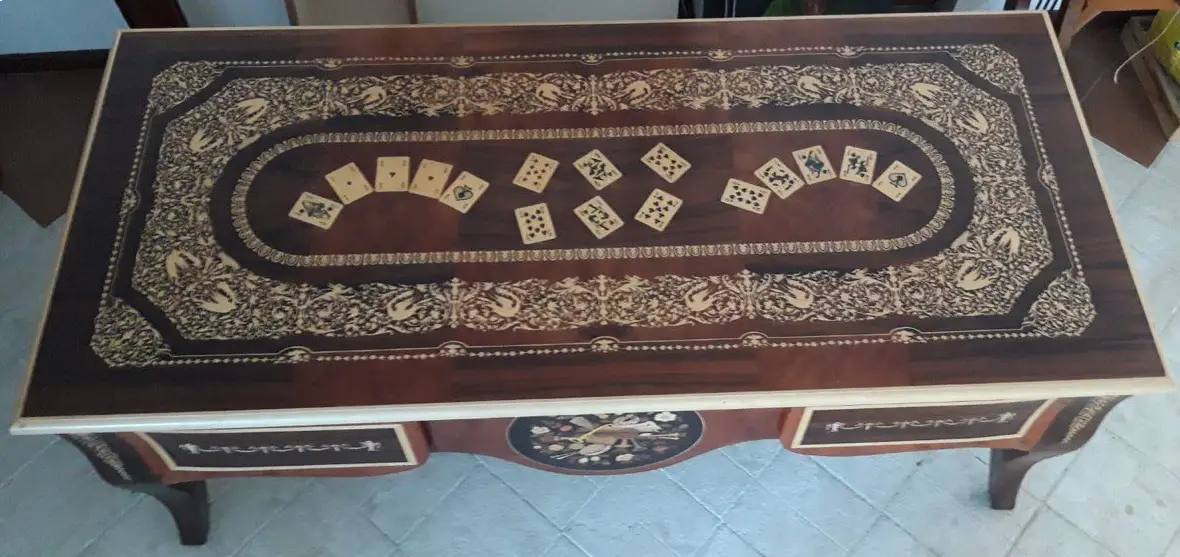 "Opera di Ay Mohsen realizzata con la tecnica dell'intarsio. Il titolo è Scrivania del poker e le tre carte e rientra nella categoria Ebanisteria Monumentale. Questa imponente scrivania monumentale rappresenta una delle opere più concettualmente audaci, raffinate e politicamente consapevoli di Aly Mohsen. In essa, la grande tradizione dell’ebanisteria artistica europea viene trasformata in strumento narrativo, simbolico e apertamente critico, dando vita a un’opera che trascende la funzione d’arredo per assumere la forma di messaggio cifrato e denuncia silenziosa. Non si tratta di un semplice mobile funzionale, ma di una vera opera d’arte tridimensionale, concepita per dialogare con il potere, smascherarne i meccanismi e mettere in crisi le apparenze di legalità e correttezza. Struttura e materiali La scrivania è caratterizzata da una struttura solida ma visivamente equilibrata, con gambe sagomate e slanciate che alleggeriscono la massa monumentale del piano. L’opera è realizzata con una selezione estremamente ricercata di legni pregiati, scelti non solo per le qualità estetiche ma anche per il loro valore simbolico: Noce nazionale e noce canaletto per la struttura principale e le superfici estese, apprezzati per la profondità cromatica e la stabilità nel tempo Radica di noce per i pannelli frontali, impiegata per ottenere superfici vibranti e irregolari, metafora visiva dell’instabilità e dell’inganno Ebano ed essenze scure tinte per i fondi degli intarsi, funzionali a rafforzare il contrasto grafico Acero e acero sicomoro per gli intarsi chiari Legni naturalmente colorati (padouk, acero, mogano, rovere, radica di noce, ebano verde ed ebano nero) per la resa cromatica delle figure e delle carte da gioco Il bordo chiaro e continuo del piano richiama deliberatamente la tradizione dei grandi tavoli da gioco, incorniciando la scena e convogliando lo sguardo verso il centro narrativo. Il piano: il gioco come metafora del potere Il piano superiore costituisce il cuore concettuale dell’opera. All’interno di una ricchissima cornice ornamentale intarsiata si sviluppa una sequenza di carte da poker disposte con apparente casualità. A una prima lettura, l’immagine suggerisce una partita in corso. A uno sguardo più attento, emerge però un elemento perturbante: le combinazioni non conducono a nessun esito logico. Le mani sono incoerenti, impossibili, volutamente manipolate. Questa ambiguità è intenzionale. Il poker, simbolo di strategia, calcolo e rischio controllato, viene qui messo in relazione con il gioco delle tre carte, archetipo universale della truffa. Il messaggio è sottile ma inequivocabile: chi crede di giocare secondo regole condivise è già vittima di un inganno. Il fronte: musica, scena e finzione Il fronte della scrivania è dominato da un grande medaglione circolare intarsiato, nel quale compaiono strumenti musicali, spartiti, carte da gioco e motivi floreali. La musica diventa metafora della messa in scena del potere: un concerto apparentemente armonioso che nasconde dissonanze profonde, una composizione elegante pensata per distrarre, sedurre e confondere. Le fasce laterali, ornate con ghirlande classiche e figure stilizzate, richiamano consapevolmente il linguaggio neoclassico e ottocentesco, rafforzando l’idea di ordine, autorità e rispettabilità — un ordine solo apparente. Storia dell’opera e valore simbolico La scrivania fu commissionata da un importante uomo d’affari egiziano, figura di enorme peso economico internazionale. Aly Mohsen, che conosceva il committente fin dall’infanzia, aveva fondati motivi per ritenere che questi operasse in realtà come prestanome per i servizi segreti statunitensi, coinvolto in operazioni economiche e finanziarie condotte in Italia. Al momento della consegna, Mohsen si recò personalmente nell’ufficio del committente e ne spiegò il significato con parole che rivelano la natura profonda dell’opera: «Questa scrivania serve per avvisare chi si siede di fronte a te: lui pensa di giocare a poker, mentre tu stai giocando al gioco delle tre carte.» Un’accusa di straordinaria eleganza, affidata non al linguaggio diretto ma alla forza simbolica dell’intarsio. Il committente, comprendendo perfettamente il messaggio, si sentì apertamente insultato e rifiutò l’opera. La scrivania rimase così nelle mani dell’artista. Rimasta nella disponibilità dell’artista, la Scrivania del Poker e del Gioco delle Tre Carte possiede oggi un valore ulteriormente accresciuto. È un pezzo unico, documentato, con una storia forte, verificabile e di straordinaria attrattiva per il collezionismo internazionale. L’opera unisce: alta ebanisteria artistica contenuto politico e narrativo qualità museale dimensioni monumentali provenienza diretta dall’artista Per collezionisti evoluti e investitori in mobili d’arte, arti decorative e opere concettuali, questa scrivania rappresenta un caso raro di arredo che diventa linguaggio critico, capace di dialogare con design, scultura e arte contemporanea, e destinato a una progressiva valorizzazione culturale e patrimoniale. In Aly Mohsen, il legno non è mai solo materia: diventa linguaggio, accusa, ironia e memoria. E questa scrivania ne è una delle dichiarazioni più lucide e implacabili."