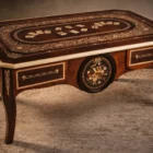 "Opera di Ay Mohsen realizzata con la tecnica dell'intarsio. Il titolo è Scrivania del poker e le tre carte e rientra nella categoria Ebanisteria Monumentale. Questa imponente scrivania monumentale rappresenta una delle opere più concettualmente audaci, raffinate e politicamente consapevoli di Aly Mohsen. In essa, la grande tradizione dell’ebanisteria artistica europea viene trasformata in strumento narrativo, simbolico e apertamente critico, dando vita a un’opera che trascende la funzione d’arredo per assumere la forma di messaggio cifrato e denuncia silenziosa. Non si tratta di un semplice mobile funzionale, ma di una vera opera d’arte tridimensionale, concepita per dialogare con il potere, smascherarne i meccanismi e mettere in crisi le apparenze di legalità e correttezza. Struttura e materiali La scrivania è caratterizzata da una struttura solida ma visivamente equilibrata, con gambe sagomate e slanciate che alleggeriscono la massa monumentale del piano. L’opera è realizzata con una selezione estremamente ricercata di legni pregiati, scelti non solo per le qualità estetiche ma anche per il loro valore simbolico: Noce nazionale e noce canaletto per la struttura principale e le superfici estese, apprezzati per la profondità cromatica e la stabilità nel tempo Radica di noce per i pannelli frontali, impiegata per ottenere superfici vibranti e irregolari, metafora visiva dell’instabilità e dell’inganno Ebano ed essenze scure tinte per i fondi degli intarsi, funzionali a rafforzare il contrasto grafico Acero e acero sicomoro per gli intarsi chiari Legni naturalmente colorati (padouk, acero, mogano, rovere, radica di noce, ebano verde ed ebano nero) per la resa cromatica delle figure e delle carte da gioco Il bordo chiaro e continuo del piano richiama deliberatamente la tradizione dei grandi tavoli da gioco, incorniciando la scena e convogliando lo sguardo verso il centro narrativo. Il piano: il gioco come metafora del potere Il piano superiore costituisce il cuore concettuale dell’opera. All’interno di una ricchissima cornice ornamentale intarsiata si sviluppa una sequenza di carte da poker disposte con apparente casualità. A una prima lettura, l’immagine suggerisce una partita in corso. A uno sguardo più attento, emerge però un elemento perturbante: le combinazioni non conducono a nessun esito logico. Le mani sono incoerenti, impossibili, volutamente manipolate. Questa ambiguità è intenzionale. Il poker, simbolo di strategia, calcolo e rischio controllato, viene qui messo in relazione con il gioco delle tre carte, archetipo universale della truffa. Il messaggio è sottile ma inequivocabile: chi crede di giocare secondo regole condivise è già vittima di un inganno. Il fronte: musica, scena e finzione Il fronte della scrivania è dominato da un grande medaglione circolare intarsiato, nel quale compaiono strumenti musicali, spartiti, carte da gioco e motivi floreali. La musica diventa metafora della messa in scena del potere: un concerto apparentemente armonioso che nasconde dissonanze profonde, una composizione elegante pensata per distrarre, sedurre e confondere. Le fasce laterali, ornate con ghirlande classiche e figure stilizzate, richiamano consapevolmente il linguaggio neoclassico e ottocentesco, rafforzando l’idea di ordine, autorità e rispettabilità — un ordine solo apparente. Storia dell’opera e valore simbolico La scrivania fu commissionata da un importante uomo d’affari egiziano, figura di enorme peso economico internazionale. Aly Mohsen, che conosceva il committente fin dall’infanzia, aveva fondati motivi per ritenere che questi operasse in realtà come prestanome per i servizi segreti statunitensi, coinvolto in operazioni economiche e finanziarie condotte in Italia. Al momento della consegna, Mohsen si recò personalmente nell’ufficio del committente e ne spiegò il significato con parole che rivelano la natura profonda dell’opera: «Questa scrivania serve per avvisare chi si siede di fronte a te: lui pensa di giocare a poker, mentre tu stai giocando al gioco delle tre carte.» Un’accusa di straordinaria eleganza, affidata non al linguaggio diretto ma alla forza simbolica dell’intarsio. Il committente, comprendendo perfettamente il messaggio, si sentì apertamente insultato e rifiutò l’opera. La scrivania rimase così nelle mani dell’artista. Rimasta nella disponibilità dell’artista, la Scrivania del Poker e del Gioco delle Tre Carte possiede oggi un valore ulteriormente accresciuto. È un pezzo unico, documentato, con una storia forte, verificabile e di straordinaria attrattiva per il collezionismo internazionale. L’opera unisce: alta ebanisteria artistica contenuto politico e narrativo qualità museale dimensioni monumentali provenienza diretta dall’artista Per collezionisti evoluti e investitori in mobili d’arte, arti decorative e opere concettuali, questa scrivania rappresenta un caso raro di arredo che diventa linguaggio critico, capace di dialogare con design, scultura e arte contemporanea, e destinato a una progressiva valorizzazione culturale e patrimoniale. In Aly Mohsen, il legno non è mai solo materia: diventa linguaggio, accusa, ironia e memoria. E questa scrivania ne è una delle dichiarazioni più lucide e implacabili."