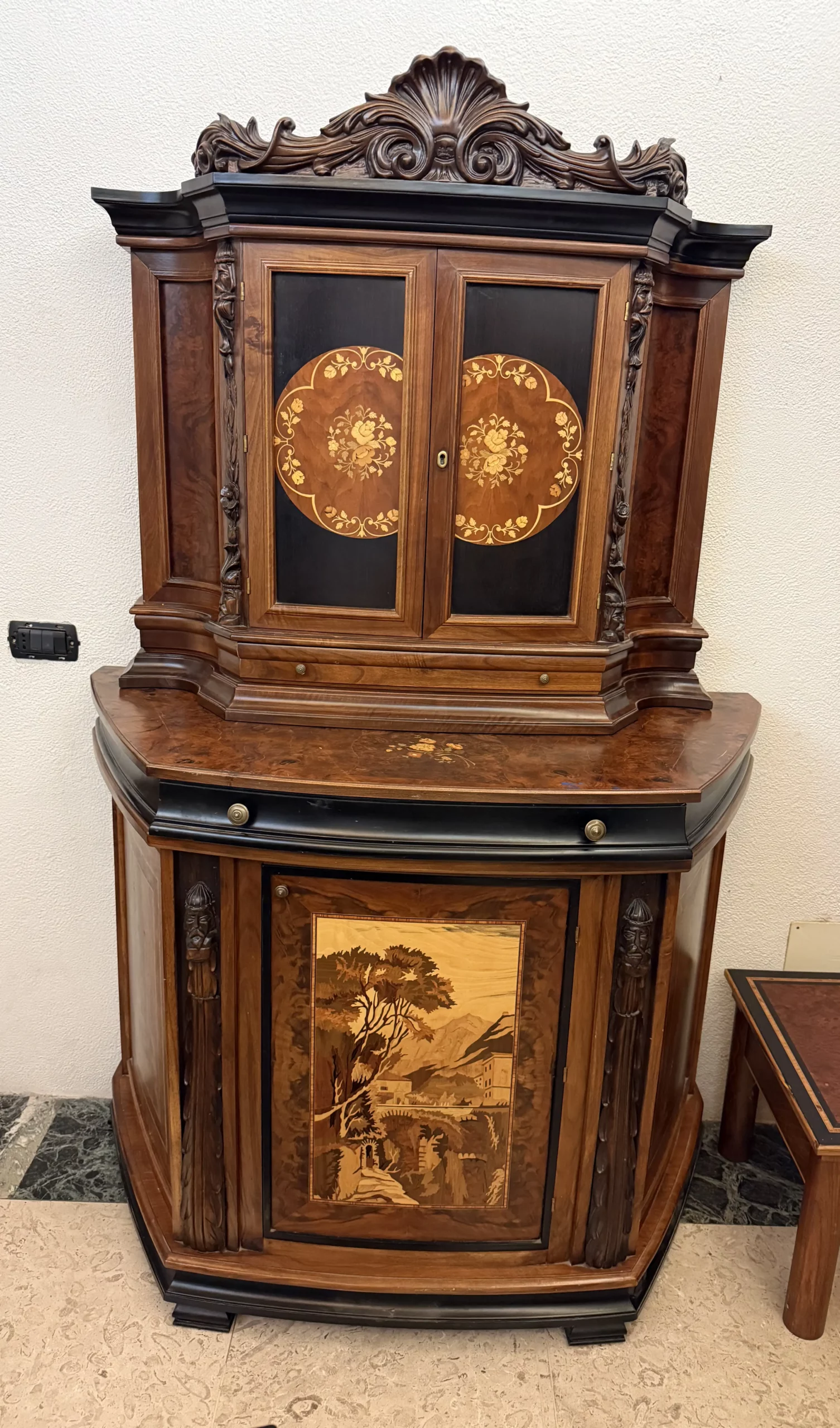"Artwork by Aly Mohsen executed in the marquetry technique. The title is Mobile del ponte vecchio (Inlaid artistic cabinet depicting the Ponte Vecchio of Ivrea) and it belongs to the category Monumental Cabinetmaking. The Credenza del Ponte Vecchio di Ivrea is an inlaid and carved artistic cabinet conceived as a unique work of high-level cabinetmaking, in which figurative tradition, authorial woodworking, and landscape reference of an identity-based nature converge. The piece presents itself as a complex three-dimensional artifact, where architectural structure, sculptural decoration, and marquetry are integrated into a coherent and unified whole. The structure is made of solid mahogany and walnut burl, used for the load-bearing elements and the main surfaces, with an alternation of compact areas and irregular grain patterns that articulate the overall volume. The sides feature hand-carved walnut sculptural elements, depicting stylized figures integrated vertically into the uprights. These carvings reinforce the cabinet’s plastic and monumental character and recall the European tradition of figurative wood carving. The upper edge and the drawer are executed in ebony, introducing a clear chromatic contrast with the warmer tones of the main structure. The upper doors are set against dark ebony grounds and enriched with polychrome floral inlays made of maple, walnut, green ebony, and cherry wood. These inlays are arranged in symmetrical compositions that establish a balance between decorative articulation and geometric order. The central element of the work is the large inlaid panel located in the lower section of the cabinet, depicting a view of the Ponte Vecchio of Ivrea. The landscape scene is constructed through a complex combination of wood species, including maple, walnut burl, light and dark walnut, and green ebony, employed to achieve tonal variation, spatial depth, and a controlled sense of light. The panel is enclosed within a rosewood frame that defines its perimeter and visually separates the image from the surrounding structure. The interior of the cabinet is fully finished and decorated, with panels featuring floral marquetry and surfaces enriched by radial geometric patterns and angular fields created through the juxtaposition of different wood species. The interior decorations are conceived as an integral part of the work and remain fully visible thanks to the presence of a glass shelf, which allows continuous visual access to the inlays when the cabinet is open. The cabinet is completed by a continuous structural framing that follows the upper and lower profiles of the piece, executed in a dark-toned wood with a uniform finish. This framing serves to visually contain the overall volume and to connect the various wooden surfaces into a coherent and unified composition."