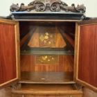 "Opera di Ay Mohsen realizzata con la tecnica dell'intarsio. Il titolo è Credenza del ponte vecchio e rientra nella categoria Ebanisteria Monumentale. La Credenza del Ponte Vecchio di Ivrea è un mobile artistico intarsiato e scolpito concepito come opera unica di alta ebanisteria, nel quale confluiscono tradizione figurativa, lavorazione lignea d’autore e riferimento paesaggistico di carattere identitario. L’opera si configura come un manufatto tridimensionale complesso, in cui struttura architettonica, decorazione plastica e apparato intarsiato concorrono alla definizione di un insieme unitario. La struttura è realizzata in mogano massello e radica di noce, utilizzati per le parti portanti e per le superfici principali, con un’alternanza di campiture compatte e venature irregolari che articolano il volume complessivo. Le fiancate presentano elementi scultorei in noce, interamente scolpiti a mano, raffiguranti figure stilizzate integrate verticalmente nei montanti, che accentuano il carattere plastico e monumentale del mobile e richiamano la tradizione europea dell’intaglio figurativo. Il bordo superiore e il cassetto sono realizzati in ebano, introducendo un contrasto cromatico netto con le essenze più calde della struttura. Le ante superiori presentano fondi scuri in ebano sui quali si sviluppano intarsi floreali policromi, eseguiti in acero, noce, ebano verde e ciliegio, organizzati in composizioni simmetriche che definiscono un equilibrio tra decorazione e ordine geometrico. Elemento centrale dell’opera è il grande pannello intarsiato posto nella parte inferiore del mobile, raffigurante una veduta del Ponte Vecchio di Ivrea. La scena paesaggistica è costruita mediante una complessa combinazione di essenze lignee, tra cui acero, radica di noce, noce chiaro e scuro ed ebano verde, utilizzate per ottenere variazioni tonali, profondità spaziale e articolazione luministica. Il riquadro è delimitato da una cornice in legno di rose che ne definisce il perimetro e ne isola visivamente l’immagine dal resto della struttura. L’interno del mobile è completamente rifinito e decorato, con pannelli intarsiati a motivi floreali e superfici arricchite da disegni geometrici a raggiera e campiture angolari ottenute mediante l’accostamento di diverse essenze lignee. Le decorazioni interne sono concepite come parte integrante dell’opera e risultano pienamente visibili grazie alla presenza di un ripiano in vetro, che consente la lettura continua degli intarsi anche a mobile aperto. La credenza si conclude con una cornice strutturale continua che accompagna i profili superiori e inferiori del mobile, realizzata in essenza scura con finitura uniforme, avente la funzione di contenere visivamente il volume complessivo e di raccordare le diverse superfici lignee in un insieme coerente e unitario."