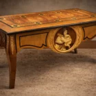 "Artwork by Aly Mohsen executed in the marquetry technique. The title is Scrivania dei fiori neri (Desk of the Black Flowers – original Italian title) and it belongs to the category Monumental Cabinetmaking. An artistic cabinetmaking desk conceived as a unique work in which functional use is integrated with an articulated decorative system of figurative character. The piece is situated within the field of contemporary authorial cabinetmaking, employing a formal language consistent with the historical tradition of wooden marquetry and hand-crafted woodworking. The tabletop is made of rosewood, with a central section in walnut burl arranged in mirrored grain along a longitudinal axis that organizes the entire composition. This central field is enclosed by a frame in walnut and white maple, within which geometric inlays in ebony, maple, and walnut are inserted. These elements are distributed in a regular manner and serve to define the visual space. The surface displays a chromatic range dominated by warm, brown tonalities, articulated through contrasts between light and dark woods. Dark-toned floral inlays are present on the tabletop, executed in ebony, maple, and padauk and arranged symmetrically along the perimeter. These elements introduce a marked chromatic contrast and contribute to the rhythmic articulation of the composition. The perimeter edge of the tabletop, executed in ebony, clearly defines the profile of the desk and reinforces its architectural reading. The legs, made of burl wood and shaped with a tapered profile, are decorated with linear white maple inlays that visually lighten their volume while maintaining formal continuity with the tabletop and side panels. The short sides feature diamond-shaped inlays with stylized floral motifs, while at the center of one of the long sides a figurative inlay depicting musical instruments is placed, composed through the juxtaposition of ebony, maple, and walnut. This inlay is enclosed by a hand-carved pearwood frame that separates the image from the structural field of the furniture. The opposite side features a figurative inlay depicting a female figure protecting a child, realized through layered woods of varying tonalities, with chiaroscuro effects achieved exclusively through chromatic variation of the materials. The composition is frontal and focused on the figures, which are set within a carved pearwood frame serving both to contain the image and to define the visual space of the figurative field. The structure includes five drawers aligned along the long side, integrated into the lower band without formal interruption, contributing to the horizontal articulation of the overall form. Light interacts with the polished surfaces and the different wood species, enhancing the depth of the grain and the legibility of the inlays, confirming the desk as a work in which cabinetmaking, figurative marquetry, and compositional organization converge into a single coherent object."