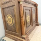 "Opera di Ay Mohsen realizzata con la tecnica dell'intarsio. Il titolo è Scrittoio delle ghirlande e rientra nella categoria Ebanisteria Monumentale. Lo Scrittoio delle Ghirlande di Aly Mohsen si configura come un mobile artistico intarsiato e scolpito, concepito come opera unica di alta ebanisteria, in cui funzione e articolazione decorativa sono integrate in una struttura di impianto architettonico. L’opera si colloca nella tradizione del mobile classico monumentale, reinterpretata attraverso una progettazione rigorosa e un controllo accurato dei materiali. La struttura portante è realizzata in noce calabrese della Sila, essenza selezionata per la compattezza, la stabilità strutturale e la profondità cromatica. I fianchi sagomati e le colonne frontali in radica di thuya introducono un ritmo plastico verticale, ulteriormente definito dalla presenza di elementi decorativi in bronzo che sottolineano i punti di raccordo e rafforzano la lettura architettonica del mobile. Elemento centrale dell’opera è il sistema di intarsi a ghirlanda, sviluppato secondo un disegno continuo e simmetrico. I profili degli intarsi sono eseguiti in acero, ebano e legno di rose, mentre i motivi floreali interni utilizzano una combinazione calibrata di acero, ebano verde, noce chiaro e noce scuro. La variazione cromatica tra le diverse essenze è costruita per gradazioni tonali e per equilibrio materico, in dialogo costante con il fondo ligneo. L’anta ribaltabile, incernierata sul fronte, rivela un interno organizzato con vani e cassetti in rovere, materiale scelto per la sua resistenza e per il contrasto visivo con le essenze esterne. Anche nella parte interna la lavorazione mantiene coerenza formale e qualità esecutiva, confermando l’unitarietà progettuale dell’opera. Il piano superiore e i bordi perimetrali sono trattati come una cornice strutturale e visiva, con funzione di contenimento dell’apparato decorativo e di definizione spaziale dell’insieme, contribuendo alla stabilità percettiva e alla chiarezza compositiva dello scrittoio."