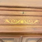 "Opera di Ay Mohsen realizzata con la tecnica dell'intarsio. Il titolo è Scrittoio delle ghirlande e rientra nella categoria Ebanisteria Monumentale. Lo Scrittoio delle Ghirlande di Aly Mohsen si configura come un mobile artistico intarsiato e scolpito, concepito come opera unica di alta ebanisteria, in cui funzione e articolazione decorativa sono integrate in una struttura di impianto architettonico. L’opera si colloca nella tradizione del mobile classico monumentale, reinterpretata attraverso una progettazione rigorosa e un controllo accurato dei materiali. La struttura portante è realizzata in noce calabrese della Sila, essenza selezionata per la compattezza, la stabilità strutturale e la profondità cromatica. I fianchi sagomati e le colonne frontali in radica di thuya introducono un ritmo plastico verticale, ulteriormente definito dalla presenza di elementi decorativi in bronzo che sottolineano i punti di raccordo e rafforzano la lettura architettonica del mobile. Elemento centrale dell’opera è il sistema di intarsi a ghirlanda, sviluppato secondo un disegno continuo e simmetrico. I profili degli intarsi sono eseguiti in acero, ebano e legno di rose, mentre i motivi floreali interni utilizzano una combinazione calibrata di acero, ebano verde, noce chiaro e noce scuro. La variazione cromatica tra le diverse essenze è costruita per gradazioni tonali e per equilibrio materico, in dialogo costante con il fondo ligneo. L’anta ribaltabile, incernierata sul fronte, rivela un interno organizzato con vani e cassetti in rovere, materiale scelto per la sua resistenza e per il contrasto visivo con le essenze esterne. Anche nella parte interna la lavorazione mantiene coerenza formale e qualità esecutiva, confermando l’unitarietà progettuale dell’opera. Il piano superiore e i bordi perimetrali sono trattati come una cornice strutturale e visiva, con funzione di contenimento dell’apparato decorativo e di definizione spaziale dell’insieme, contribuendo alla stabilità percettiva e alla chiarezza compositiva dello scrittoio."