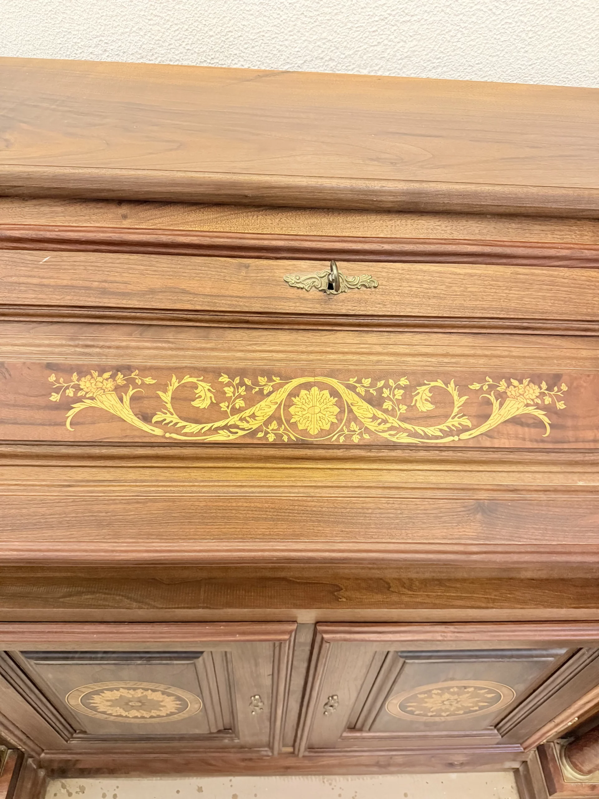 "Opera di Ay Mohsen realizzata con la tecnica dell'intarsio. Il titolo è Scrittoio delle ghirlande e rientra nella categoria Ebanisteria Monumentale. Lo Scrittoio delle Ghirlande di Aly Mohsen si configura come un mobile artistico intarsiato e scolpito, concepito come opera unica di alta ebanisteria, in cui funzione e articolazione decorativa sono integrate in una struttura di impianto architettonico. L’opera si colloca nella tradizione del mobile classico monumentale, reinterpretata attraverso una progettazione rigorosa e un controllo accurato dei materiali. La struttura portante è realizzata in noce calabrese della Sila, essenza selezionata per la compattezza, la stabilità strutturale e la profondità cromatica. I fianchi sagomati e le colonne frontali in radica di thuya introducono un ritmo plastico verticale, ulteriormente definito dalla presenza di elementi decorativi in bronzo che sottolineano i punti di raccordo e rafforzano la lettura architettonica del mobile. Elemento centrale dell’opera è il sistema di intarsi a ghirlanda, sviluppato secondo un disegno continuo e simmetrico. I profili degli intarsi sono eseguiti in acero, ebano e legno di rose, mentre i motivi floreali interni utilizzano una combinazione calibrata di acero, ebano verde, noce chiaro e noce scuro. La variazione cromatica tra le diverse essenze è costruita per gradazioni tonali e per equilibrio materico, in dialogo costante con il fondo ligneo. L’anta ribaltabile, incernierata sul fronte, rivela un interno organizzato con vani e cassetti in rovere, materiale scelto per la sua resistenza e per il contrasto visivo con le essenze esterne. Anche nella parte interna la lavorazione mantiene coerenza formale e qualità esecutiva, confermando l’unitarietà progettuale dell’opera. Il piano superiore e i bordi perimetrali sono trattati come una cornice strutturale e visiva, con funzione di contenimento dell’apparato decorativo e di definizione spaziale dell’insieme, contribuendo alla stabilità percettiva e alla chiarezza compositiva dello scrittoio."